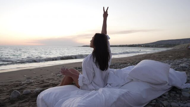 Young joyful woman showing peace gesture sitting on white soft bed admiring sunset over ocean outdoors. Wide shot of happy relaxed confident Caucasian lady enjoying leisure at dusk. Slow motion