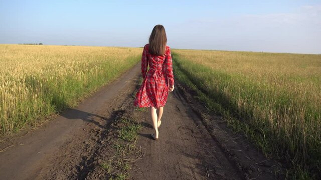 Young woman in a red dress is walking along the road along a wheat field with ripe golden spikelets, then starts running. Concept of freedom