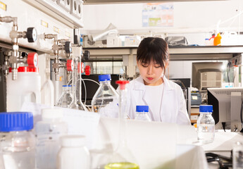 Fototapeta premium Young female Chinese student working in laboratory, writing report on results of chemical experiments in notebook