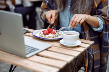 Obraz na płótnie Canvas Mature woman eats tasty toast with cut strawberries and cream near laptop and cup of coffee at table on outdoors cafe terrace closeup