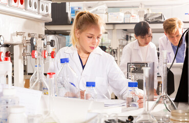 Young intelligent female student working in college laboratory, writing report on results of chemical experiments in notebook