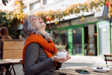 Cheerful senior lady with glasses and cup of tasty coffee laughs throwing back her head at table on...