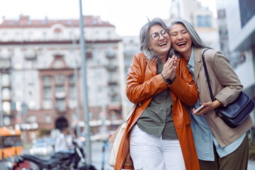 Joyful senior woman and Asian companion holding smartphone stand on modern city street on nice autumn day. Friends spend time together