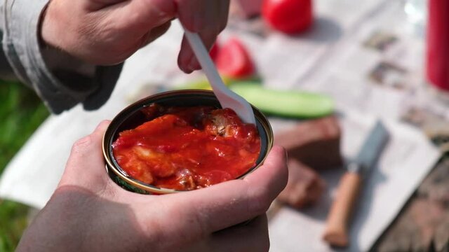 Picnic in nature. A man takes out canned fish in tomato sauce with a fork. In the background an impromptu table with vegetables and bread. Fast food, delicious snack.