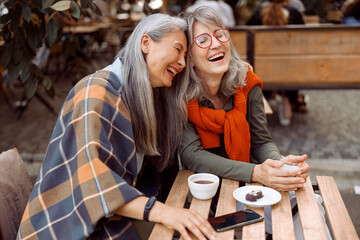Pretty senior Asian woman puts head on laughing friend shoulder resting together at table in street...