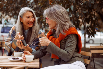 Cheerful senior Asian lady shows phone to grey haired friend at small table with coffee and candies...