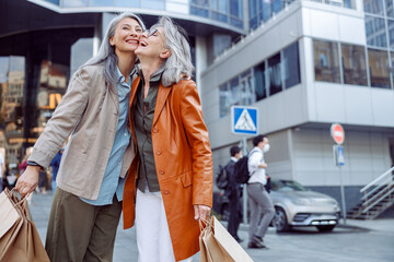 Joyful senior woman with paper shopping bags hug Asian lady on modern city street on autumn day. Friends spend time together