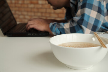 Thai male worker busy working with laptop, use chopsticks to hastily eat instant noodles during office lunch's break, because quick, tasty, and cheap. Over time Asian fast food, unhealthy lifestyle.