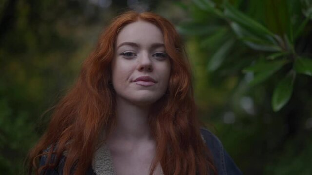 Beautiful redhead woman with long flowing hair walks through a stunning garden in autumn. A romantic and cinematic scene of a red haired woman wearing a long evening dress and warm jacket