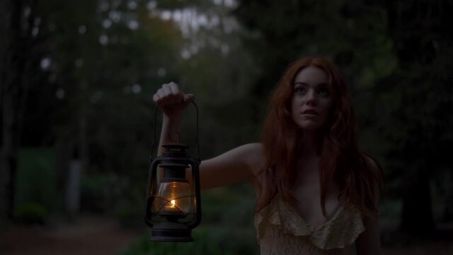 Beautiful redhead walks through a forrest in the evening holding a lantern, searching for her true love while feeling cautious and nervous and afraid while searching by candlelight, Halloween feeling