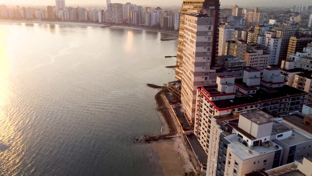 Long drone flight along with the highrise buildings of the stunning Sao Vincente shoreline. Sunset with beautiful light.  Some people exercising on the pavement below.