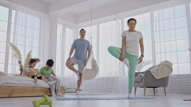 Joint family leisure activity, parents practicing yoga while their preadolescent kids playing on bed. Athletic father and mother exercising together in light spacious domestic room during free time