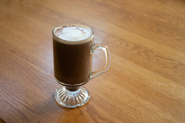 cup full of fresh cappuccino with cinnamon on the top of foam angled to the side from a distance sitting on a wooden kitchen table for fall