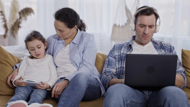 Addicted father in headphones busy on laptop while left to themselves mother and little daughter using smartphone and communicating with each other. Modern family spending free time online