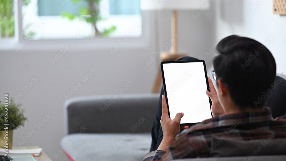 Poster back view casual man sitting on comfortable sofa and using digital tablet.
