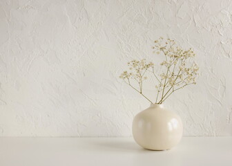 Modern beige ceramic vase with dry  flowers on white table near a textured white concrete wall....