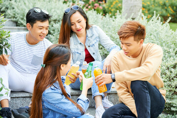 Cheerful young people resting outdoors and clinking bottles of refreshing cold drinks