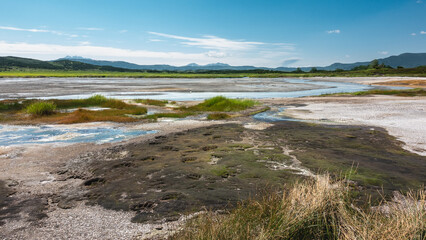 The river of hot springs flows in the caldera of an extinct volcano. The grass grows on stony soil. Bear paw prints are visible on the moss. A mountain range against the blue sky. Kamchatka. Uzon
