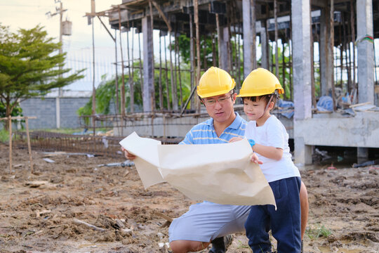 Smiling Happy Asian Dad And Son Wearing Yellow Construction Helmet Or Safety Hard Hat Standing In Front Of Construction Sit, Father And Son Working On Building A New House, Home Building Project 