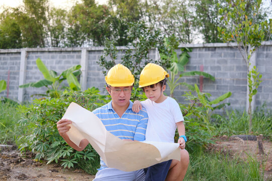 Smiling happy Asian Dad and son wearing yellow construction helmet or safety hard hat standing in front of construction sit, Father and son working on building a new house, Home building Project 