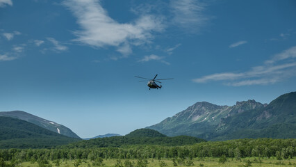 Fototapeta premium The helicopter descends into the valley. There is green grass and trees in the meadow. A picturesque mountain range against the blue sky. Kamchatka.