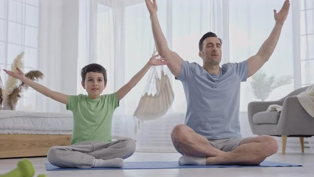Handsome man training yoga meditation together with preteen son sitting on mat in light domestic room. Caring dad and preadolescent boy doing yoga exersices during joint training at home