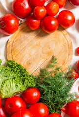 Round cutting wooden board, around lies a mountain of red tomatoes, leaves of greenery, dill. Vertical
