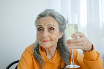Beautiful old grandmother with grey hair and face with wrinkles sitting at the table at home on window background with glass of champagne, mother's day, happy retirement