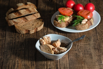 On a wooden background, there are ready-made sandwiches with cod liver, tomato and greens, canned fish are lined in a dark plate. 