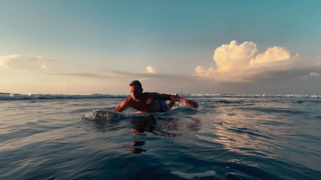 Young slender shirtless surfer guy in shorts swims by, rowing with his hands, lying on a surfboard in the ocean at sunset, clouds, blue sky, waves, slow motion.