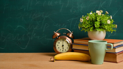 Books, vintage alarm clock, banana, mug, plant and mockup space over green blackboard