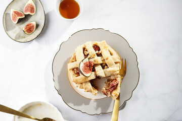 A sweet fig tart pastry on a sophisticated plate, two golden dessert spoons, honey, cut figs on a marble surface, top view