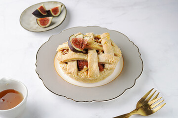 A sweet fig tart pastry on a sophisticated plate, two golden dessert spoons, honey, cut figs on a marble surface