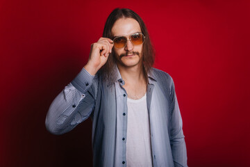 A man with long hair in a plaid shirt and white T-shirt, in blue jeans. The guy with the mustache and dark glasses is dressed in the style of the 70s and 80s. Simple working guy.