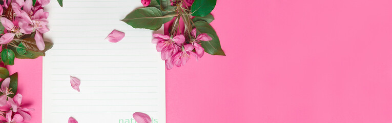 Top view of blank sheet of notebook and branches of apple blossoms on pink background. Copy space. Banner