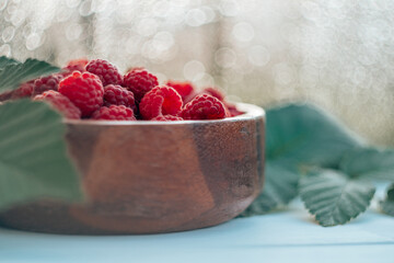 Partially blurred wooden round bowl full of ripe red juicy raspberries on bokeh background with raspberry bush leaves