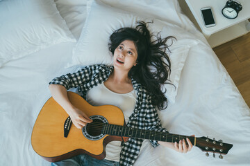view from top happy asian female musician is singing and playing an acoustic guitar while lying on bed at leisure in the bedroom at home. - Powered by Adobe
