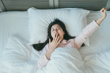 Fototapeta na wymiar high angle shot sleepy asian woman wearing pajamas is lifting her arm and covering her mouth while yawning after waking up in the early morning on bed at home.
