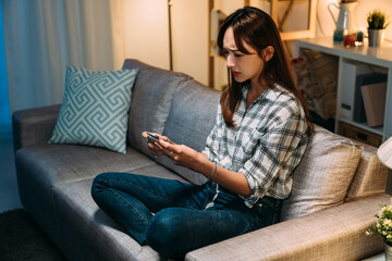 Fototapeta premium upset asian young woman playing mobile games is frowning at the phone in hands feeling unhappy about losing on the sofa in the living room at home.