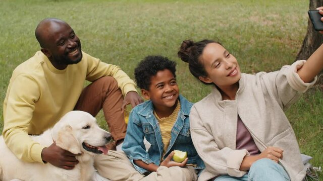 Cheerful Afro-American family sitting in park with golden retriever dog and taking selfie with smartphone while having summer picnic