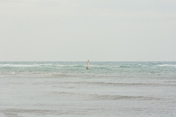 Naklejka premium Windsurfing in the open sea alone. Pastel gray autumn seascape with small waves and a man with a sail on the horizon. The concept of sports, privacy, recreation. Landscape minimalism, neutral color