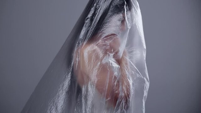 Female trafficking. Social outcast. Woman rights. Isolation problem. Profile of insecure hopeless girl captive covered with plastic wrap in gray out of focus copy space background.