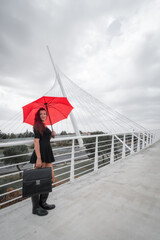 White female business executive smiling with work briefcase, 40s long reddish brunette hair in black dress, wellies and open red umbrella on modern city bridge. Toledo, Spain. Vertical view