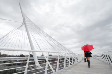 White female business executive smiling with business briefcase, 40s long reddish hair brunette with black dress, wellies and red umbrella walking with back to camera on modern city bridge. Toledo, Sp