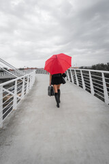 White female business executive smiling with business briefcase, 40s long reddish hair brunette with black dress, wellies and red umbrella walking with back to camera on modern city bridge. Toledo, Sp