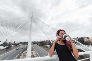 White female business executive drinking coffee and talking on the phone with work briefcase in her 40s with long reddish brown hair in black dress, on modern city bridge over freeway with moving cars