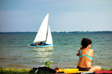 Fototapeta premium A young woman sits in a swimsuit on the beach by the lake and sunbathes, in the background is a sailing yacht with a crew