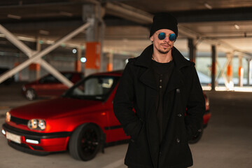 Fototapeta na wymiar Stylish handsome young man model with fashion sunglasses and a hat in a vintage black coat near a red car in the city at the parking