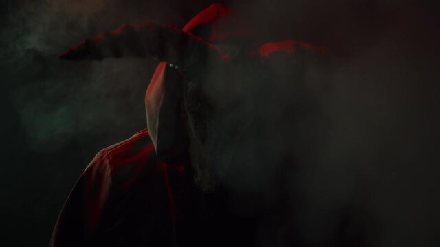 Devil with cow skull head making rite in smoke. Demon with horns conjuring close-up. Evil imp wearing black robe with hood using witchcraft in darkness and fog. Halloween and full moon.