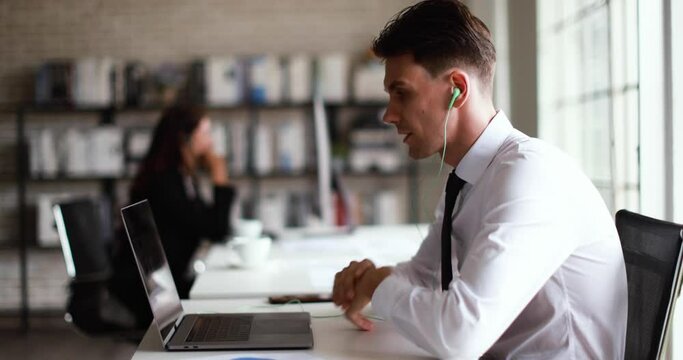 Smart young businessman sitting at desk and talking with team in online conference in office with another businesswoman speaking with other internet meeting session in blur background.